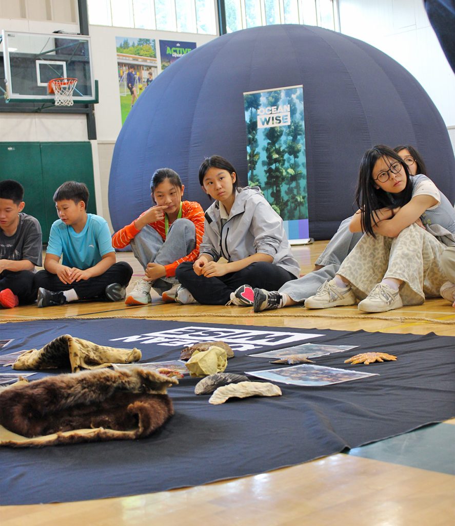 A group of children sit on the floor with marine artifacts spread out in front of them. Behind them is the Ocean Wise Sea Dome